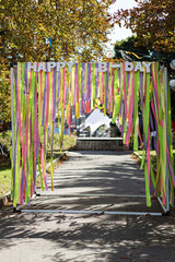 Beautiful colorful ribbons and happy birthday words hanging in sunny park at street food festival, photo zone. Bright rainbow decorations at street food market or birthday. Summer picnic