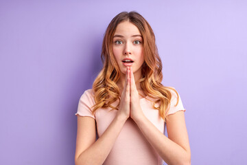 Attractive blonde girl in casual clothes prays for wellness of family, keeps palms pressed together in praying gesture, dreaming. Studio shot, purple background. Mock up copy space