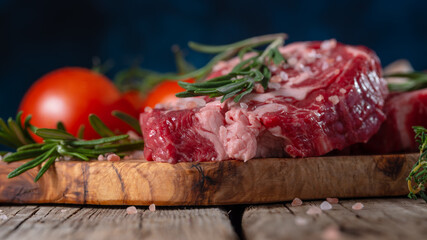 In the photo, raw meat for cooking steaks. The meat lies on a cutting board. Above is a sprig of dill. In the background are tomatoes. Wooden table. There are no people in the photo. Close-up.