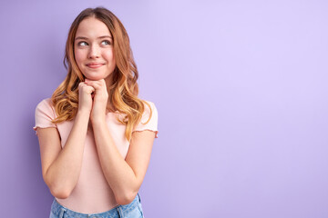 Young blonde female with wavy hair looking at side with dreamy face, calm and smiling, in casual clothes, isolated on purple background. portrait