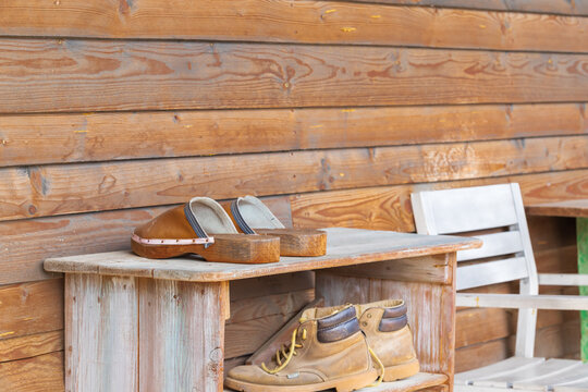 Old Wooden Shoebox Near A Wooden Cottage On A Farm. The Shoe Box Has Old Clogs, Rubber Boots And Shoes.