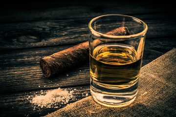 Glass of tequila and piece of cloth with salt and cuban cigar on an old wooden table. Close up view, focus on the cuban cigar