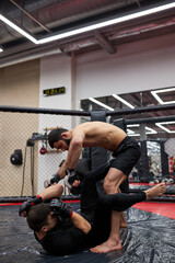 Fototapeta na wymiar Concentrated muscular boxer wearing gloves and shorts kicking competitor in gym lying on floor. Two young professional sportsmen combating in fight without rules. Concept of extreme sport.