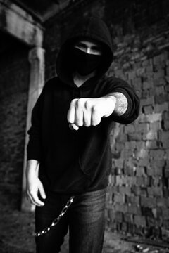 Young handsome criminal gangster model man in a black mask against covid coronavirus. Protest in the hood or hoodie near the orange brick wall. Strength, aggression, and ready for thefight. urban life