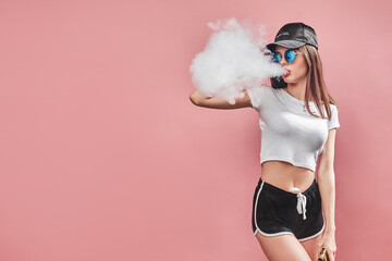 Seductive glamour young woman standing and vaping on pink background