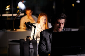 Obraz na płótnie Canvas Determined businessman concentrated hard on difficult computer task working late in dark office looking worried, in formal wear, colleagues working on laptop in the background. copy space