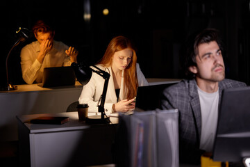 Team of managers office workers working on project very late, using computer. Hardworking people in formal wear engaged in work, thinking, brainstorming, focus on redhead female with smartphone