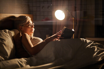 Beautiful female in pajamas and eyeglasses lying on bed reading book, alone at night. Caucasian short haired lady in bedroom, charming cute woman in room lighted by lamp