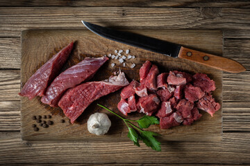 fresh raw beef meat cut into pieces in the process of cooking lies on a wooden cutting board with a kitchen knife, herbs and spices against the background of old planks. top view