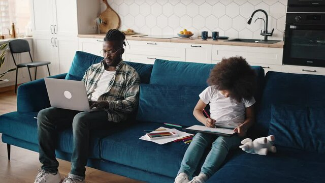African american man working on laptop on sofa, his cute little daughter drawing with crayons nearby, sitting at home