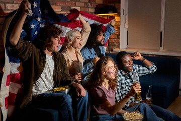 Cheerful american friends soccer fans gathered together at home in dark room watching on tv...
