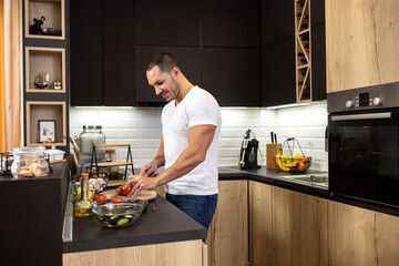 Smiling muscular man in the kitchen slicing a tomato with a chef knife as he prepares salad