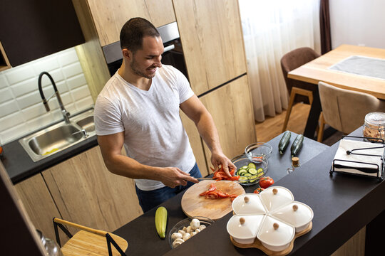 Handsome man slicing a tomato in his kitchen with a knife, plant based food