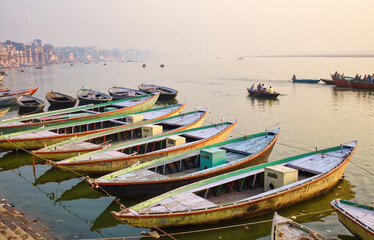Fototapeta na wymiar Varanasi, India : Bunch of old wooden colorful boats docked in the bay of Ganges river bank during sunset sunrise against foggy weather. The holy place for hindu religion.