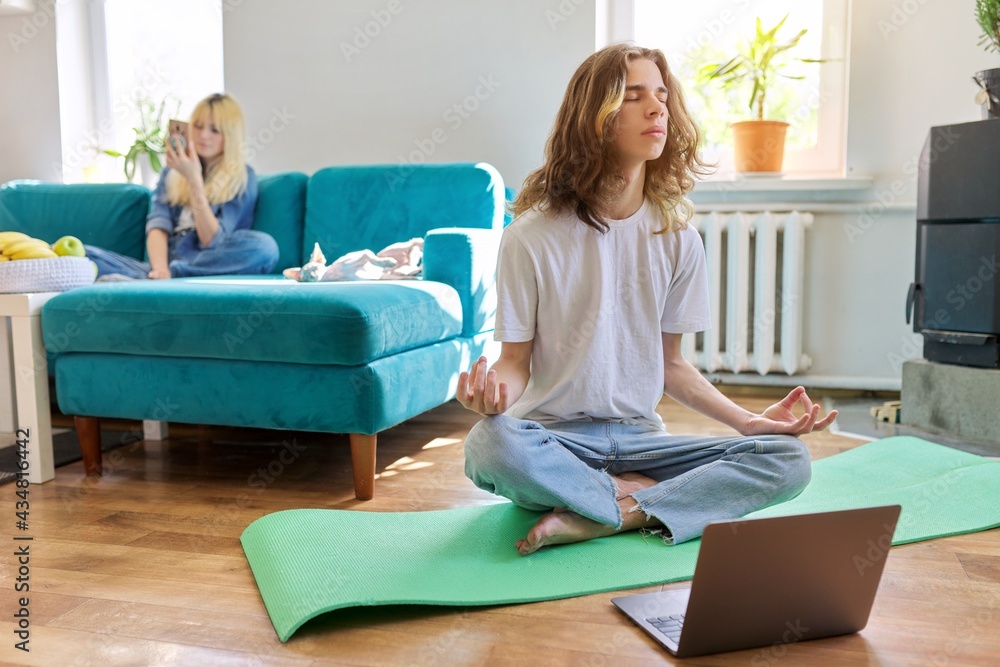 Wall mural Guy teenager sitting in lotus position on yoga mat at home on floor with laptop