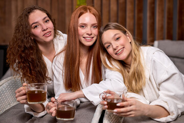 Three young ladies clothed bathrobe lying on deckchairs, talking and drinking tea, caucasian charming women having rest, leisure time, posing looking at camera. Spa procedures. beauty concept