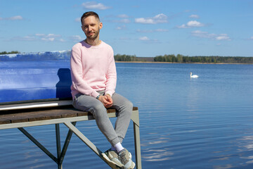 A young handsome guy sits on a pier with a boat near the lake. Floating swan in the background