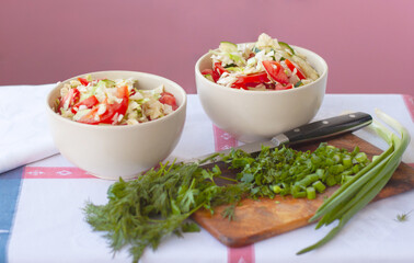 Two light bowls with a salad of fresh vegetables are on the table covered with a tablecloth, next to a cutting board, a knife, parsley and onions