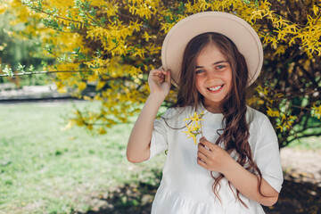 Pretty little smiling girl holding flowers under blooming forsythia bush in spring garden.