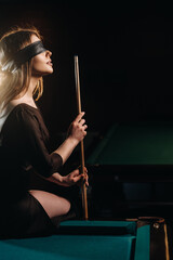 A girl with a blindfold and a cue in her hands is sitting on a table in a billiard club.Russian...