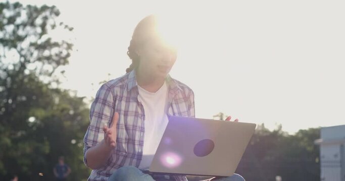 Cheerful young woman in colorful shirt talks gesticulating at videocall on laptop sitting at back sunlight in spring park closeup