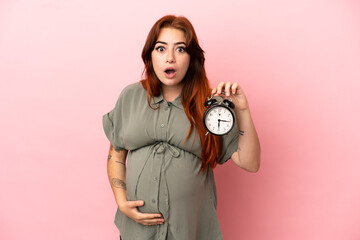 Young redhead caucasian woman isolated on pink background pregnant and holding clock with surprised...