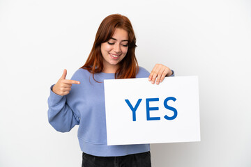 Young redhead woman isolated on white background holding a placard with text YES and  pointing it