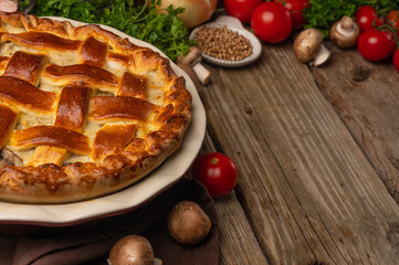 On a wooden table is a delicious baked pie with mushrooms. Appetizing golden brown crust. In the foreground are mushrooms, tomatoes. In the background, spices, herbs, tomatoes.