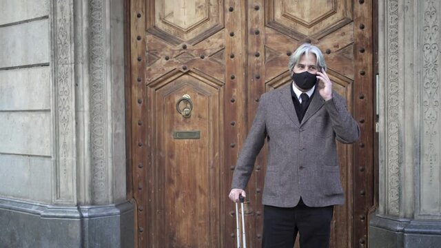 Senior Businessman with face mask and travel luggage calling with cell phone in front of wooden hotel gate