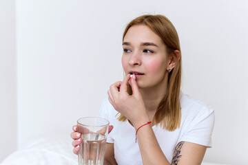 Young woman with glass of water taking sleeping pills and not feeling well in bed, copy space. Using medical drug at home. Vitamin or supplement