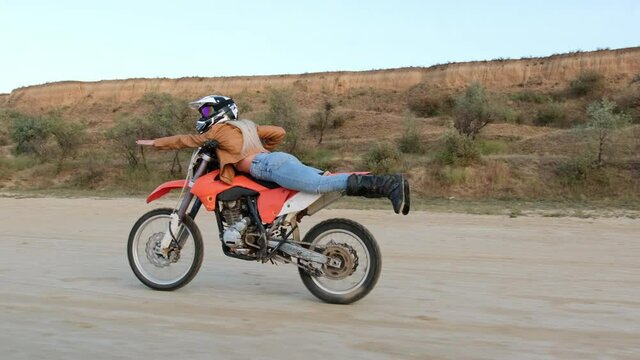 Brave young woman does stunts riding red motorbike on ground road in countryside area against dry grass and thickets field
