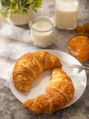 In the photo there are two croissants on a white plate with an appetizing crust. In the background is orange jam in a vase and a glass of milk. light background, contrasting orange and beige colors.