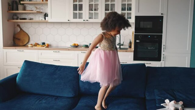 Happy childhood. Adorable african american girl wearing dress jumping on sofa at home, laughing and having fun