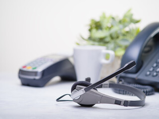 Office dialing - telephone, headphones, headset. Light and dark gray tones. In the background is a white cup and a flower in a pot. Light pastel colors. Bright daylight.