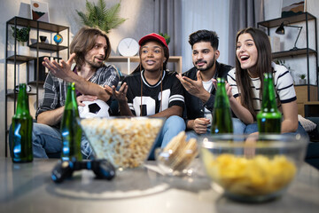 Four multiracial friends sitting on couch and discussing soccer match that watching on TV. Happy men and women eating snack and drinking beer during free time at home.