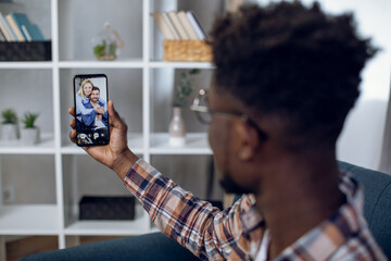 Happy african man having video call with caucasian friends using modern smartphone. Young guy staying at home during pandemic and enjoying distance communication.