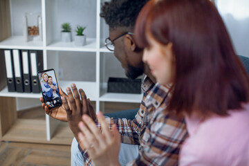 Back view of multi ethnic couple waving hands during video conversation on smartphone with friends. People using modern technology while staying at home.