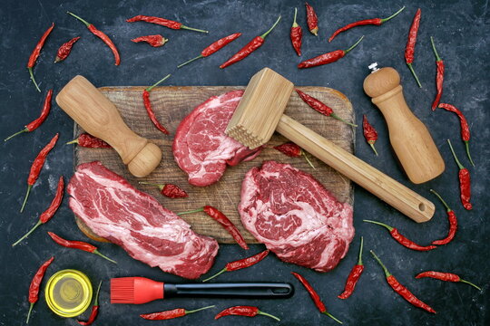 Beef Steaks for Grilling Or Frying, Overhead View. Marbled Raw Loin Beef Steaks, Wooden Hammer, Salt and Pepper Mill On Cutting Board. Raw Striploin Marbled Beef Steaks on Black Background, Top View.