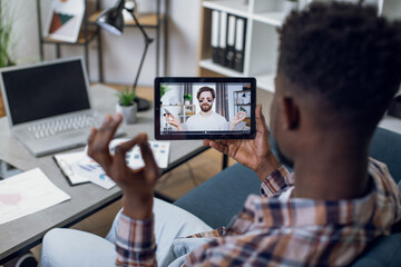 View from shoulder of african man talking with caucasian psychologist during video call on tablet. Online consultation during pandemic. People and technology concept.