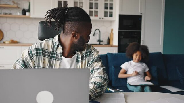 Happy african american dad working on laptop computer, smiling to little daughter, enjoying remote work at home