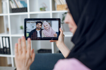 Young woman in hijab having video conversation with muslim friends from home. Pretty lady holding digital tablet and waving hand for greeting.