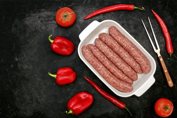 Sausages For BBQ Grilling or Frying in White Ceramic Dish And Red Peppers on Grungy Black Background, Top View. Raw Beef or Lamb Barbecue Sausages in Natural Casing in White Tray, Overhead View.