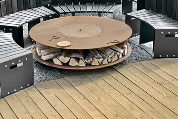 Round Patio Fire Pit Steel Table For Outdoor Leisure Party On The Wood Deck Terrace. Iron Rounded Fire Pit With Grill Top On Backyard Party Place. Grill Appliance And Fireplace On The Back Yard Lawn.