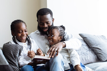 Father reads a book to his two daughters on sofa whilst they laugh