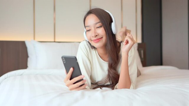 Beautiful happy smiling Thai woman lying on bed with headphones listening to music while holding mobile phone in hotel room