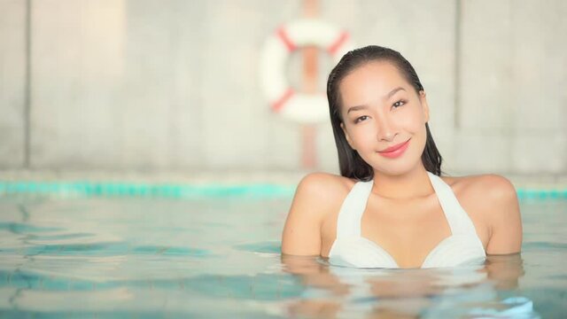 Attractive cheerful Thai young woman immersed in pool water takes bath with lifebuoy in background