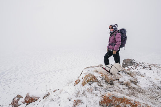 Portrait of tourist female standing on the edge of cliff over frozen sea with white dog. Winter hike in Siberia