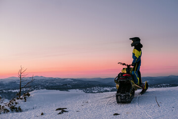 Snowmobile rider standing on top of mountain during sunset with atmospheric landscape view