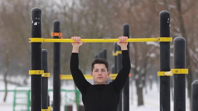 A young man pulls himself up on a horizontal bar on the street in winter. 