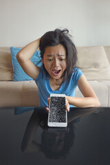 Angry woman sitting alone on couch in living room at home looking at smartphone with crashed screen.
